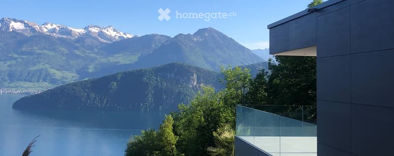 Modern building with a glass balcony overlooking a lake and snow-capped mountains.