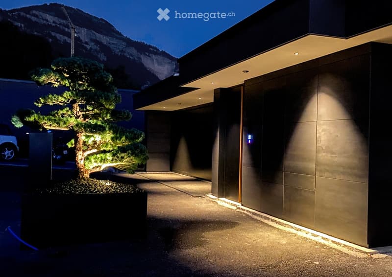Modern house exterior at night with a well-lit bonsai tree in a planter, sleek architecture, and a mountain view in the background.