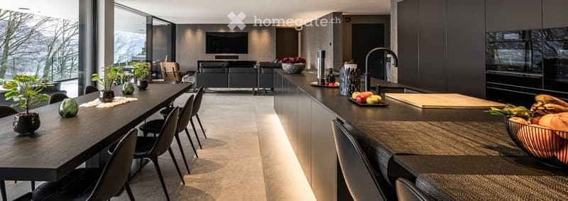 Modern open-plan kitchen and dining area with dark wood surfaces, sleek black chairs, a long dining table, a modern sofa, and a large window offering a scenic mountain view.