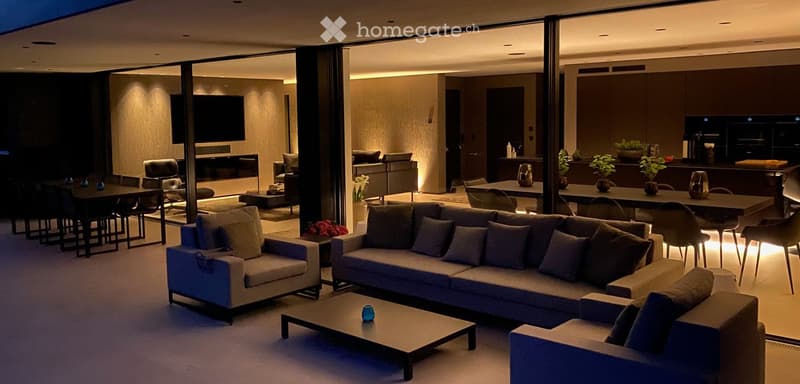 Modern living and dining area with a large sectional sofa, coffee table, dining table with chairs, fireplace, and a kitchen island with bar stools and potted plants.