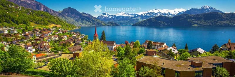Scenic view of a Swiss village with a lake and snow-capped mountains in the background, surrounded by lush green trees and houses.
