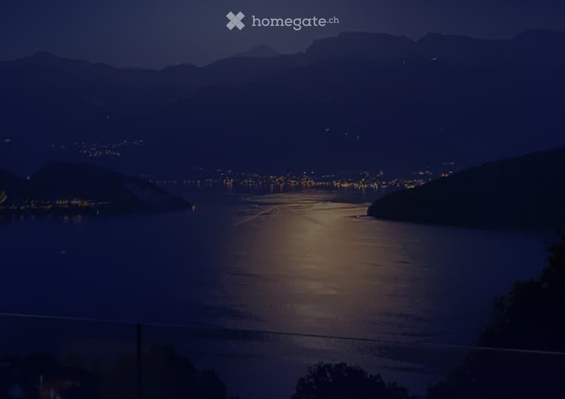 Nighttime view of a lake with city lights across the water, surrounded by dark hills and mountains.