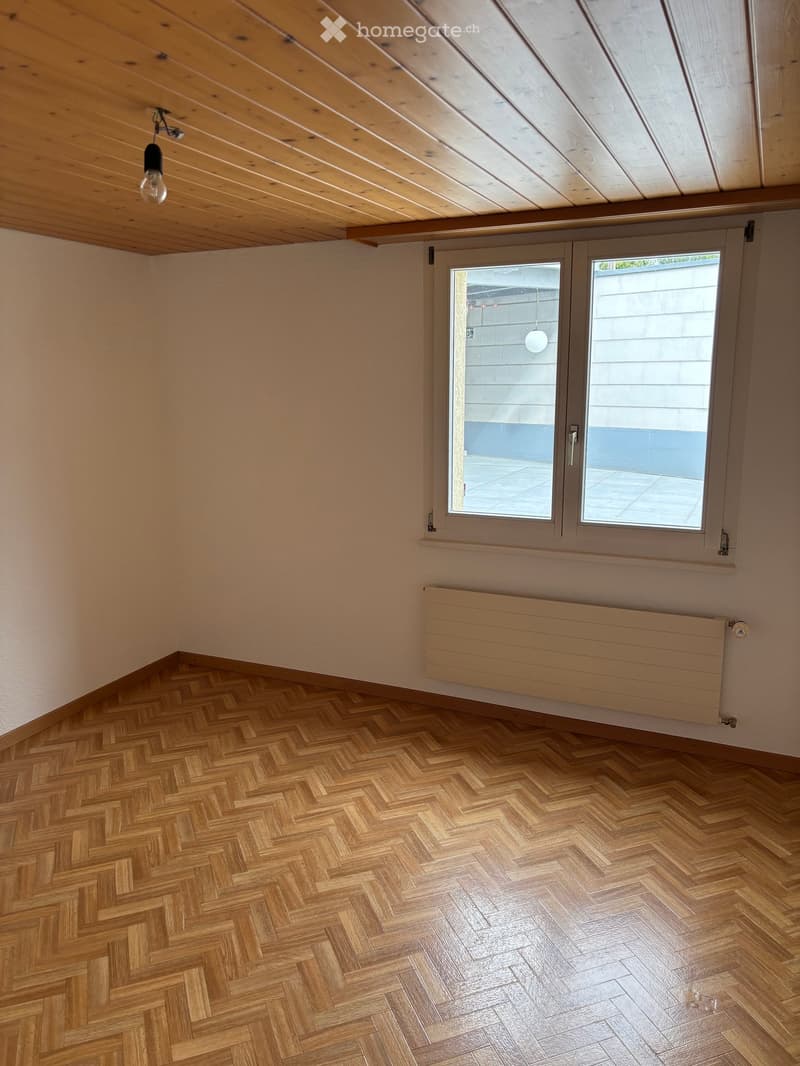 Empty room with herringbone wooden flooring, exposed wooden ceiling beams, large window with sliding doors, and a radiator below the window.