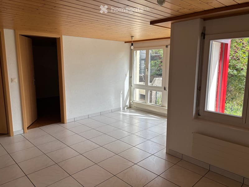 Empty room with tiled floor, white walls, wooden ceiling, and large windows offering a view of greenery outside.