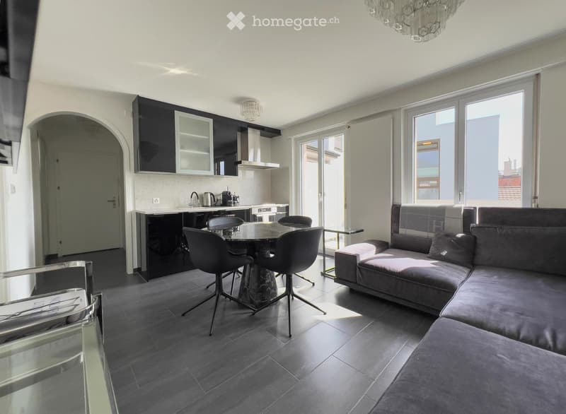 Modern open-plan living and dining area with grey sectional sofa, glass coffee table, dining chairs, and a sleek kitchen with black cabinets and stainless steel appliances.