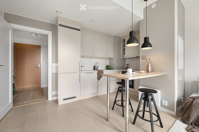 Modern kitchen with light grey cabinets, a wooden countertop, and two black hanging pendant lights. The space includes a sink, a small table with two stools, and a vase with dried flowers on the counter.