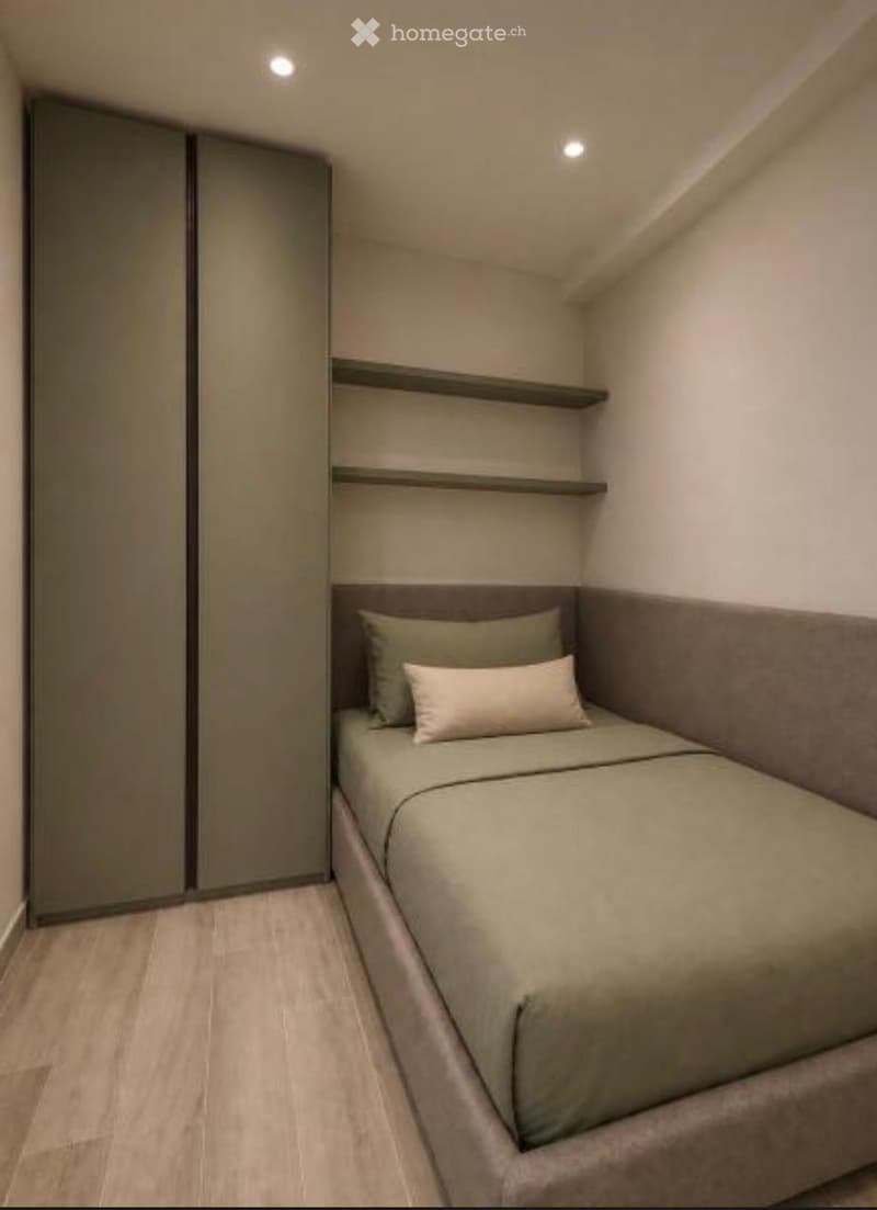 Modern, compact bedroom with a single bed, built-in wardrobe, and shelves. The bed features a headboard and is neatly made with pillows and a duvet.