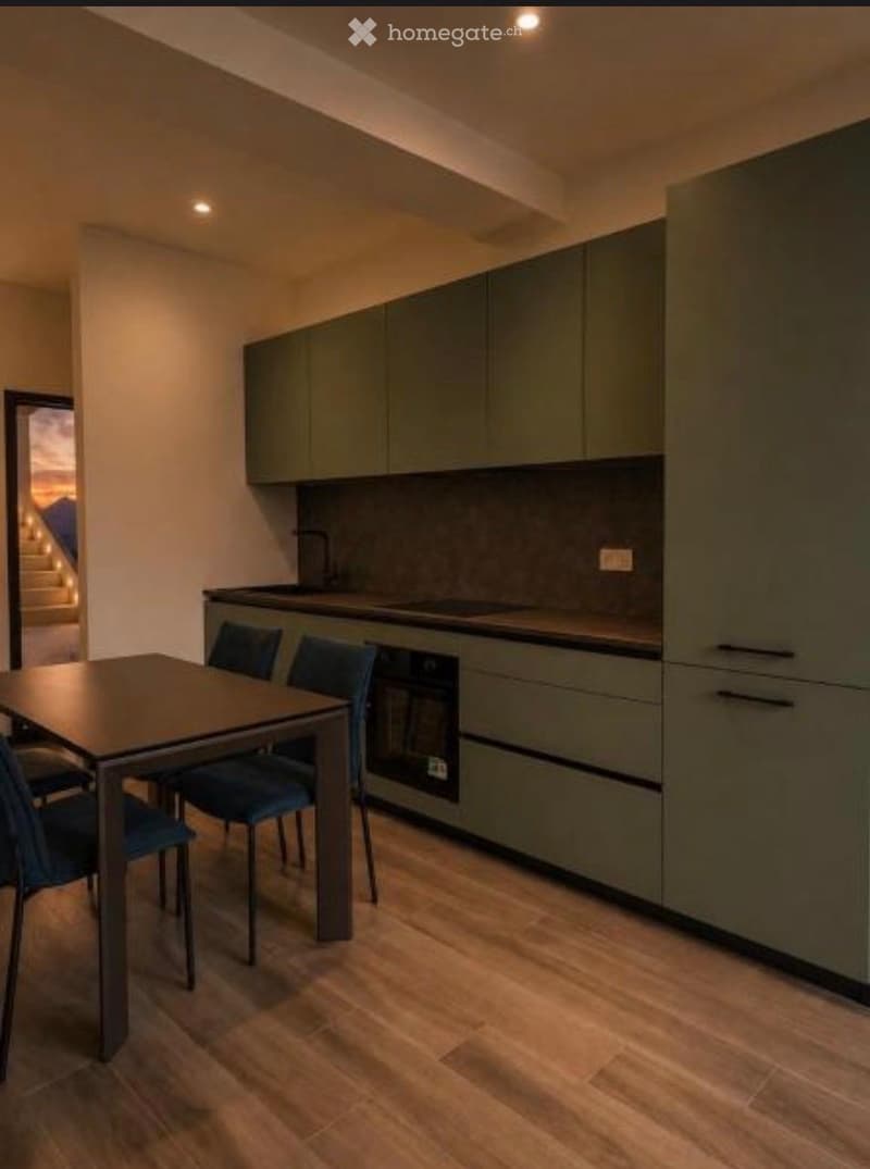 Modern kitchen with fitted green cabinets, a dark countertop, and a dining area featuring a table and chairs.