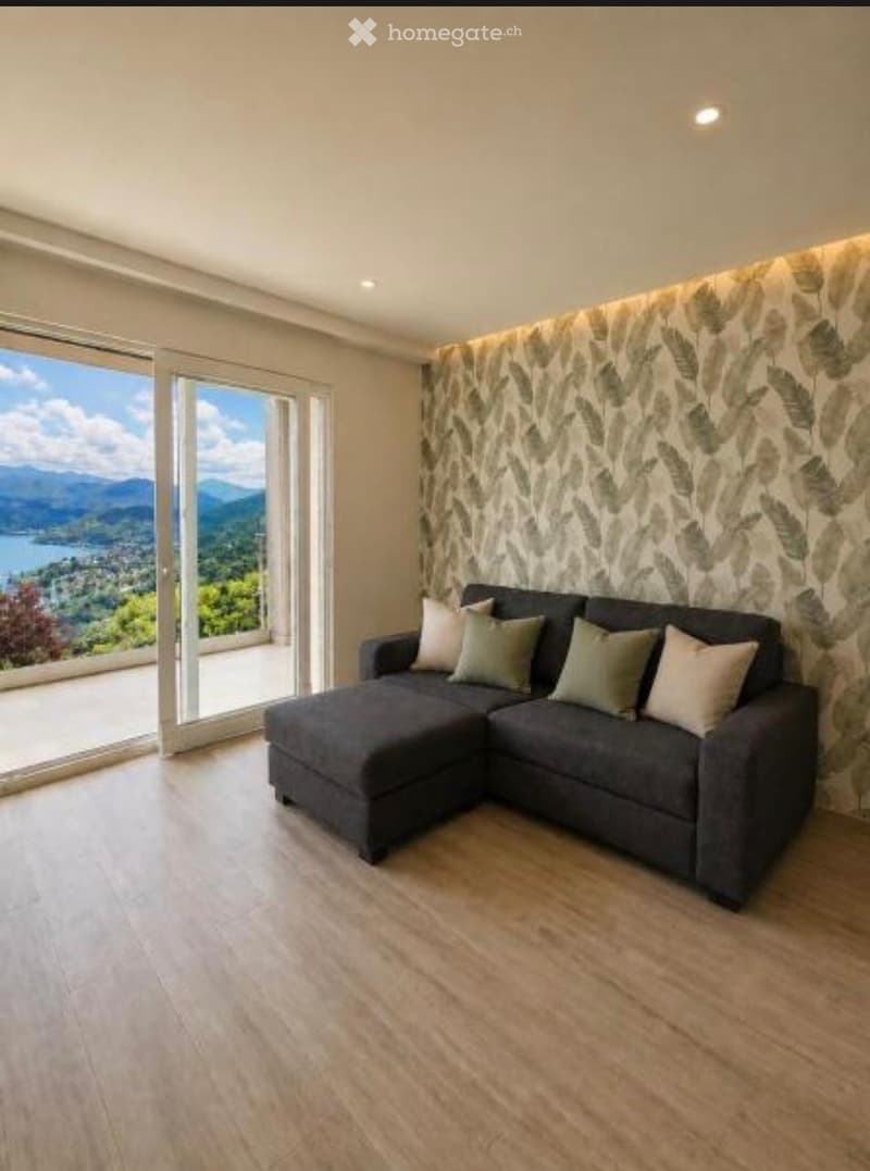 Modern living room with a grey sectional sofa, multiple cushions, large windows offering a scenic mountain and lake view, and light wooden flooring.