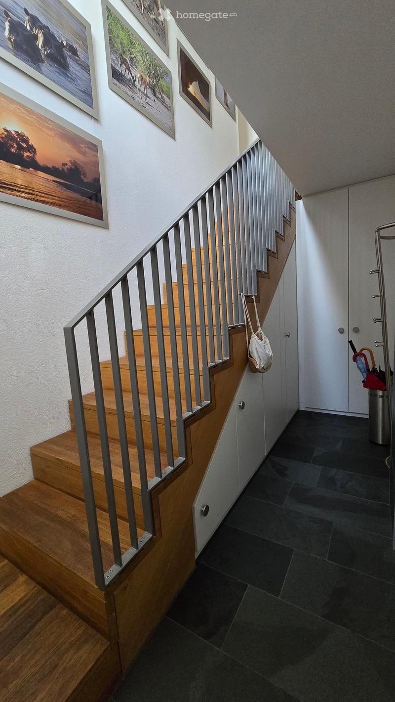 Modern hallway with wooden staircase, metal railing, and framed artwork on the wall. Dark tiled floor and white walls.