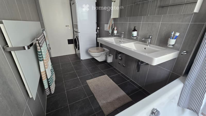 Modern bathroom with dark tiled walls and floor, double sink, bathtub, toilet, and a small rug in front of the sink.