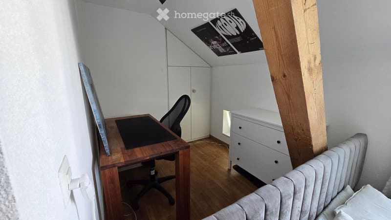 A modern, furnished room with a wooden desk, ergonomic chair, white cabinet, and radiator. The room features wooden flooring and white walls, with a slanted ceiling and a poster on the wall.