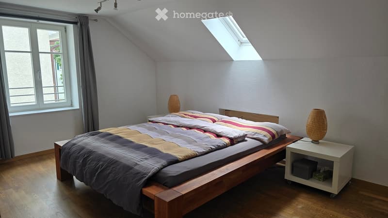 Cozy attic bedroom with wooden bed, nightstand, table lamp, and large window with curtain. Slanted ceiling and skylight add natural light.