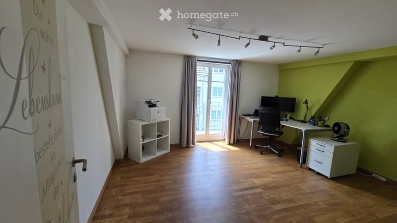 Office space with wooden flooring, large window with curtains, desk with chair, computer screen, printer on cabinet, and small storage unit.