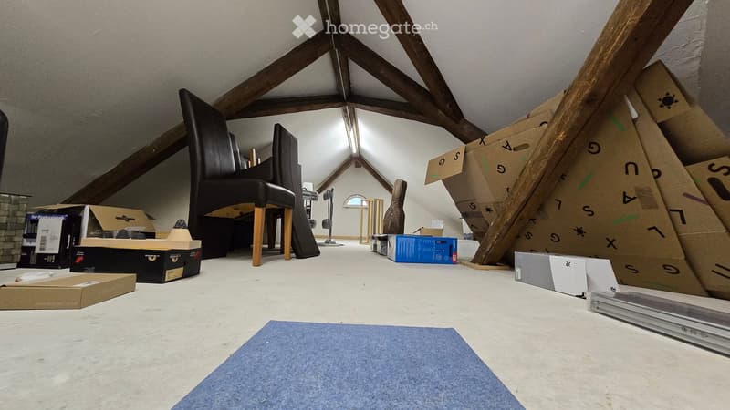 An attic space with exposed wooden beams, a black leather chair, a guitar case, and several cardboard boxes stacked around the room.