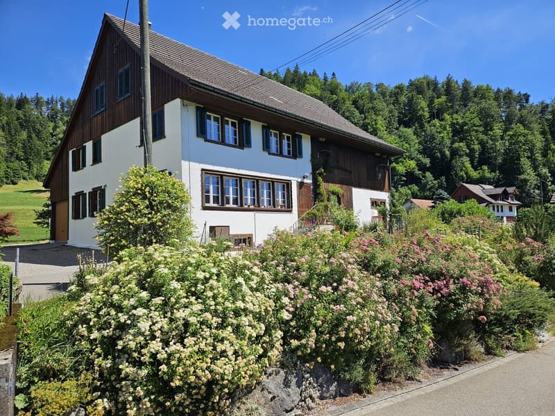 Two-story house with white walls, wooden trim, multiple windows, and a sloped roof, surrounded by lush greenery and flowers.