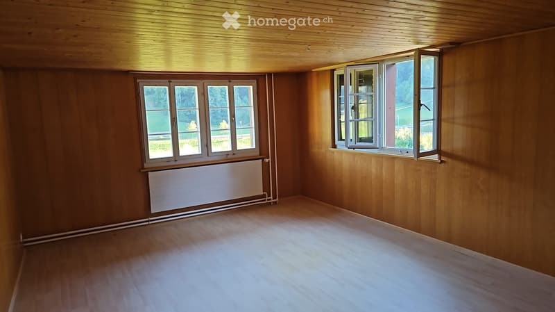 Empty room with wooden walls, wooden floor, and large windows with white frames. A radiator is visible on one wall.