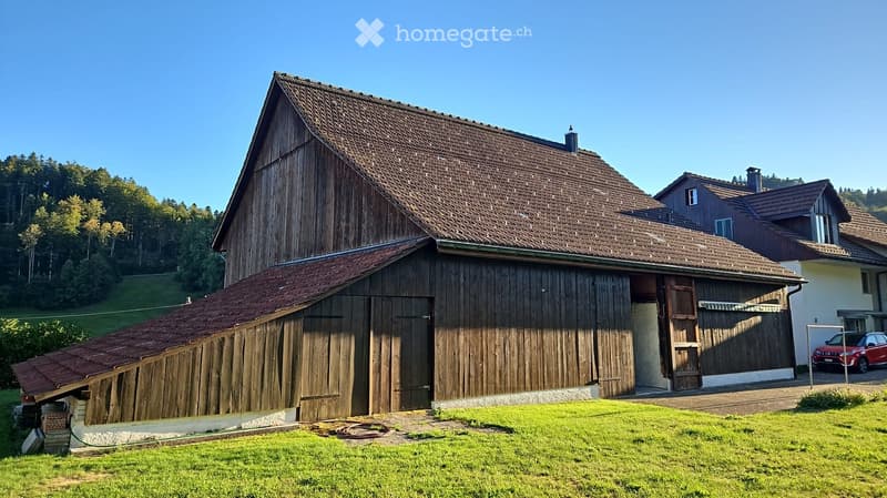 A large wooden barn with a steep, tiled roof and a smaller attached structure, surrounded by a grassy area and trees in the background.