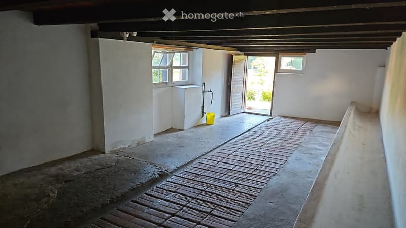 Empty room with white walls, concrete floor, and a small window. There is a doorway leading outside and a small utility area with a sink and a yellow bucket.