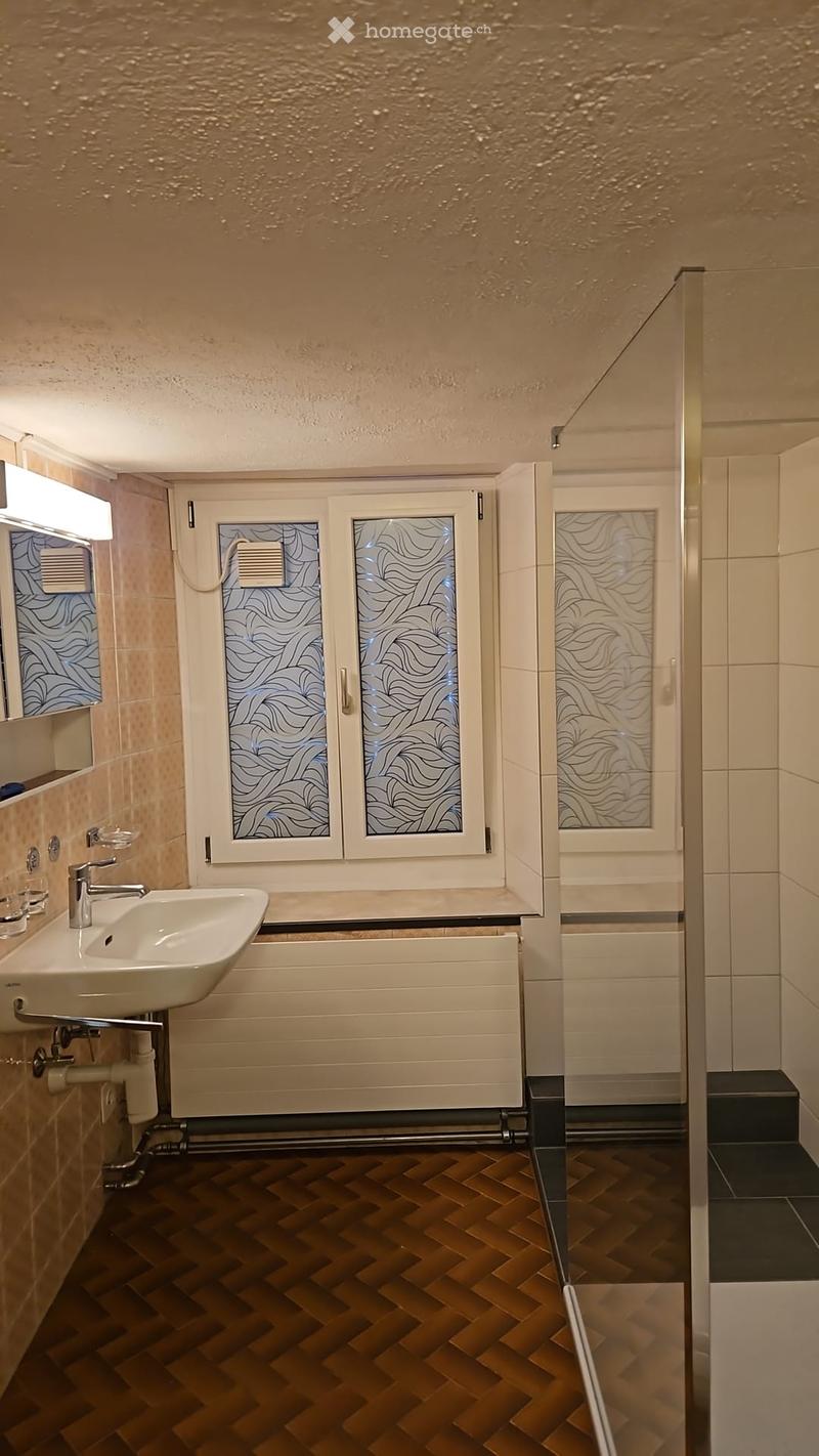 Modern bathroom with a white sink, chrome faucet, mirrored cabinet, and a glass-enclosed shower area. The floor features a herringbone pattern in warm wood tones.