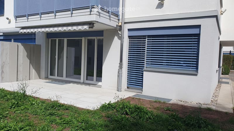 Modern two-story building with white exterior, large sliding glass doors, covered balcony, and neatly landscaped garden area.