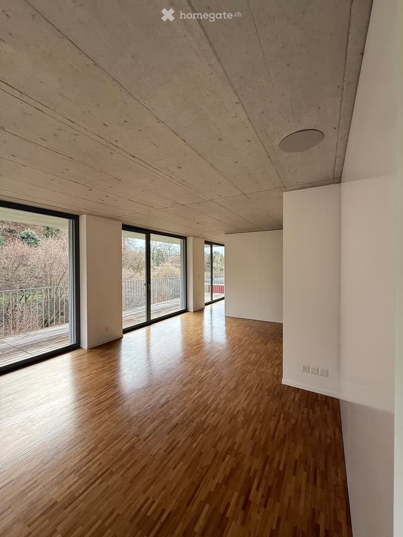 Empty room with wooden flooring, white walls, large windows, and a slanted wooden ceiling.