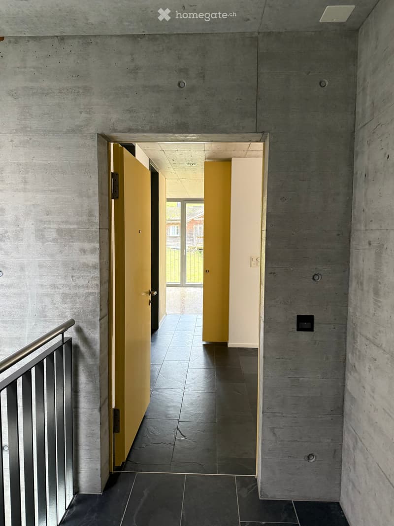 Modern hallway with concrete walls, dark tiled floor, and yellow doors leading to other rooms. A metal railing is visible on the left side.