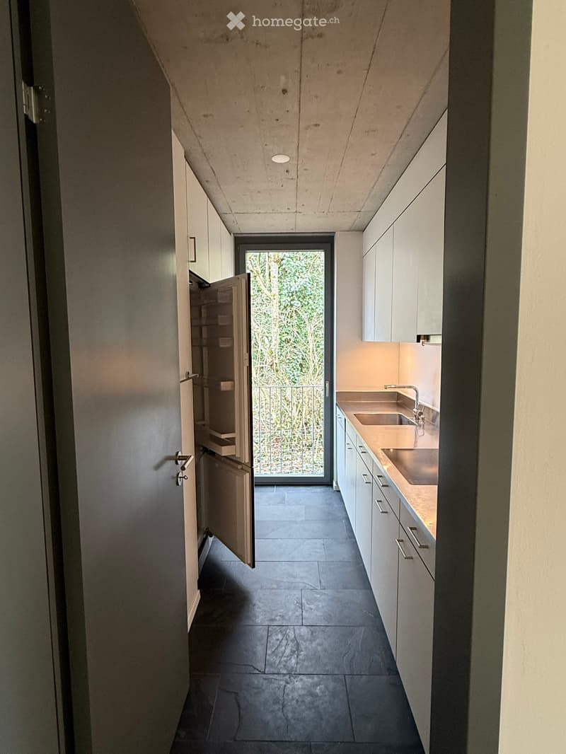Modern kitchen with white cabinets, dark tiled floor, and a sink with a faucet. A sliding glass door leads to a balcony with greenery visible outside.