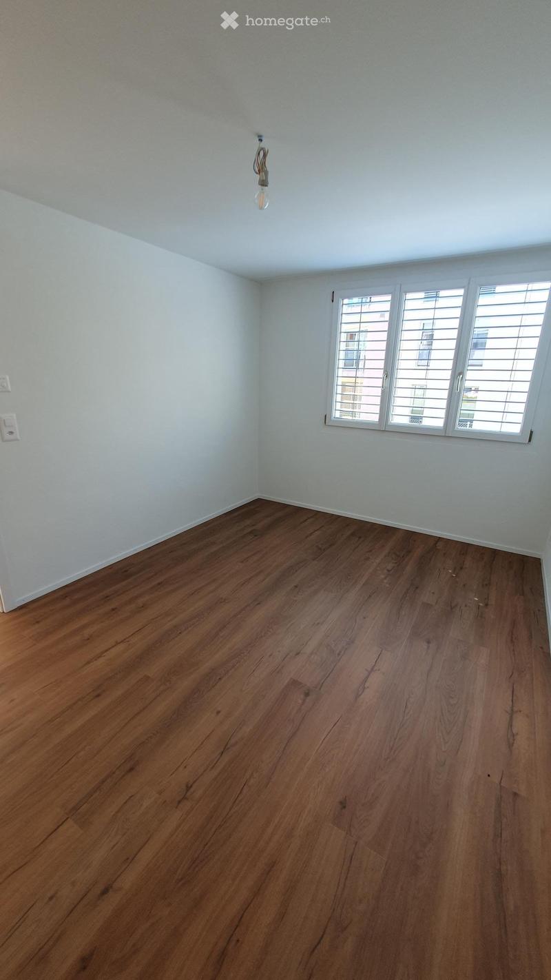 Empty room with wooden flooring and white walls, featuring a window with blinds and a single light fixture on the ceiling.