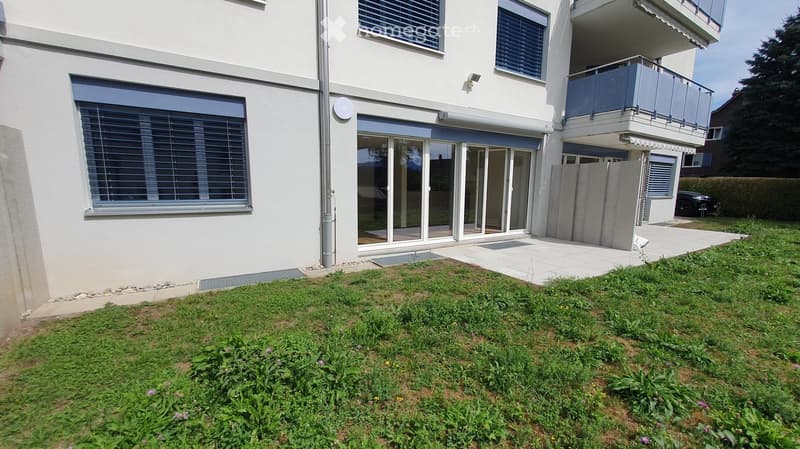Modern two-story building with white exterior, large windows, sliding glass doors, and a balcony on the upper level. The ground floor has a covered entrance area and a small garden with grass and flowers.