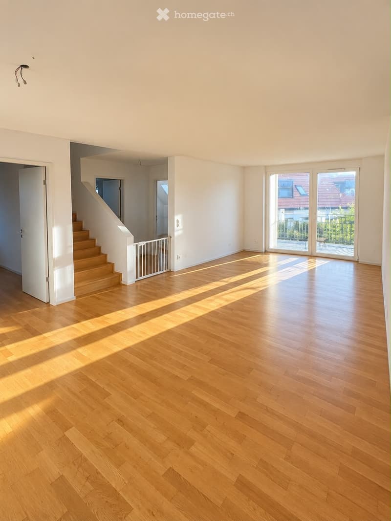 Empty room with wooden flooring, white walls, and large windows letting in natural light. A staircase leads to another level, and a baby gate is visible near the stairs.