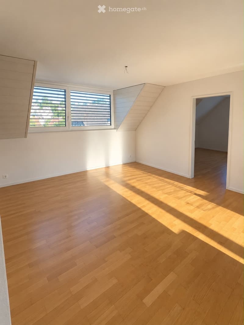 Empty room with wooden flooring, white walls, and large windows with blinds. Sunlight streams in, casting shadows on the floor.