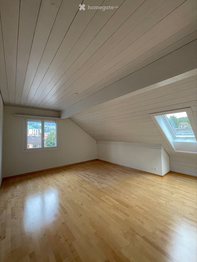 Empty attic room with wooden flooring, white walls, and skylight windows, featuring a slanted wooden ceiling.
