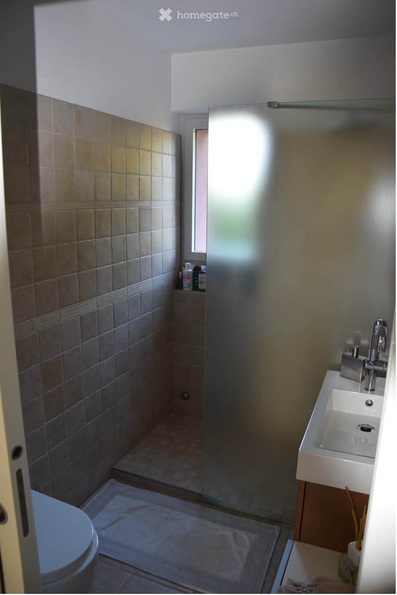 Modern bathroom with a white sink, chrome faucet, toilet, tiled walls, and a small shelf with toiletries.