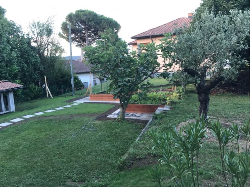 Well-maintained garden with a lawn, trees, and a pathway leading to a raised planter area. The garden is surrounded by residential buildings and features a mix of greenery and paved areas.