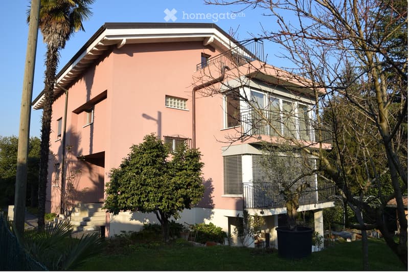 Two-story pink house with white trim, multiple windows, and a balcony on the upper level. Surrounded by trees and greenery.