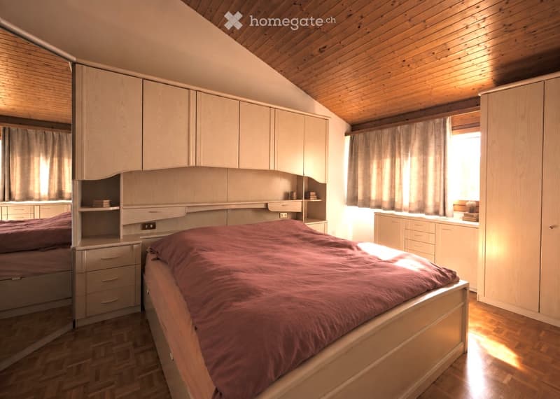 Cozy bedroom with wooden ceiling, large bed with red bedding, built-in wardrobes, nightstand, and a mirror. Natural light filters through the window with curtains.