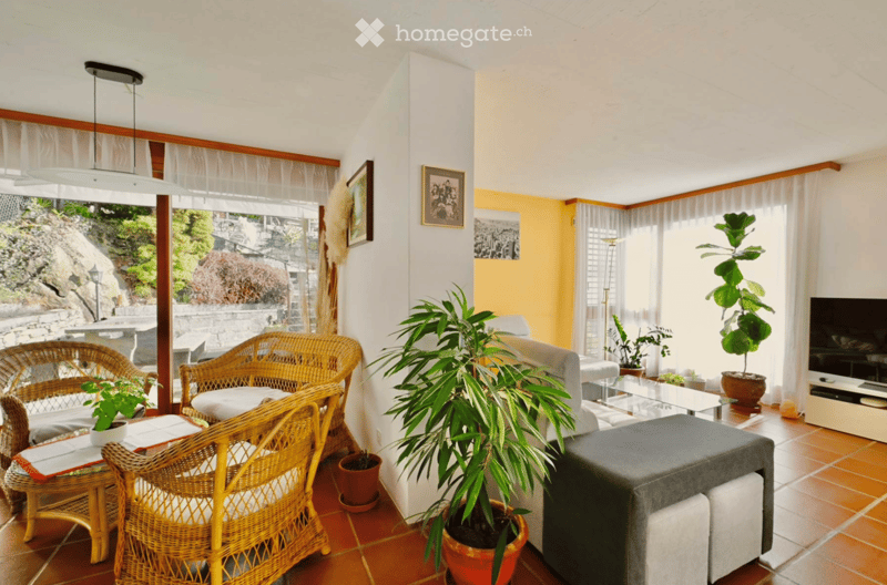 Bright living room with large windows, wicker chairs, sofa, coffee table, ottoman, potted plants, and television.