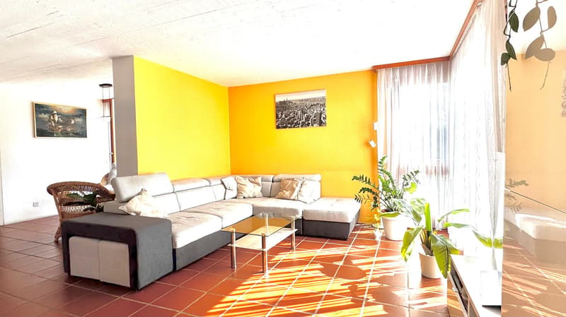 Bright living room with a grey sectional sofa, coffee table, ottoman, and potted plants. The room features a yellow accent wall, terracotta tile flooring, and large windows with sheer curtains.