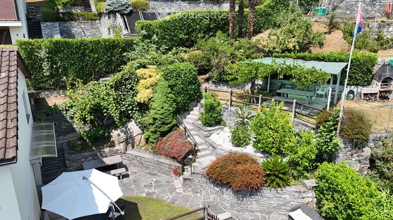 A well-maintained garden with stone pathways, neatly trimmed hedges, and various plants. Features include outdoor seating areas, a barbecue, and a flagpole.