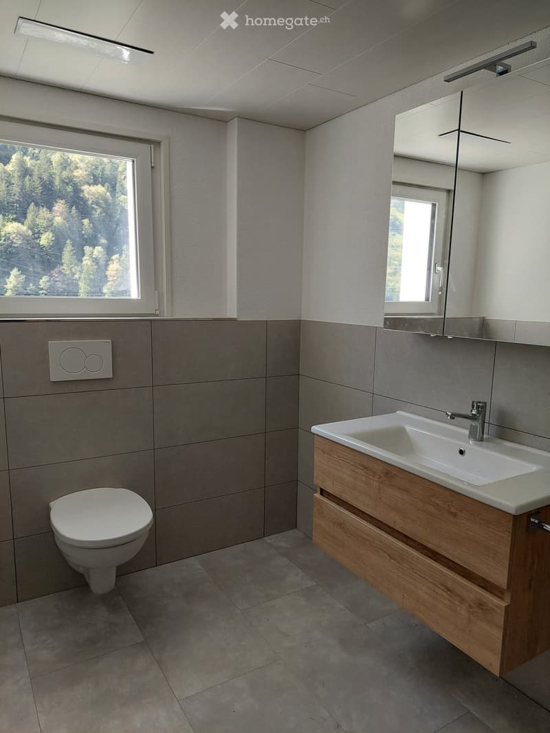 Modern bathroom with a toilet, double sink, wooden cabinet, and large mirror. Grey tiled walls and floor, with a window offering a view of trees.