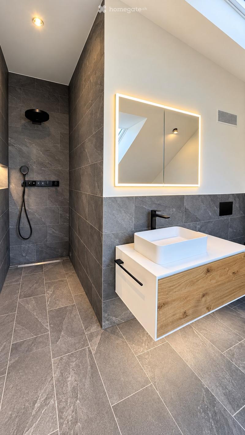 Modern bathroom with grey tiled walls and floor, a white sink on a wooden vanity, a large illuminated mirror, and a separate shower area with a handheld showerhead.