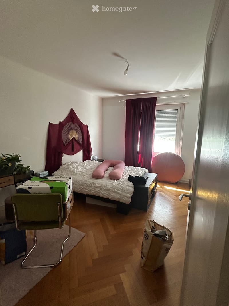 Bedroom with wooden flooring, a bed with pillows, a chair, a large orange ball, and a curtained window.