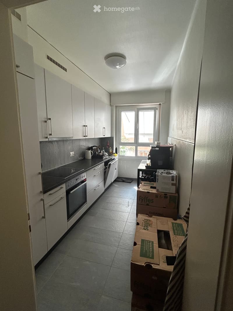 A small kitchen with white cabinets, a black countertop, and a window overlooking a balcony. There are several cardboard boxes stacked near the window, and a coffee maker is on the counter.