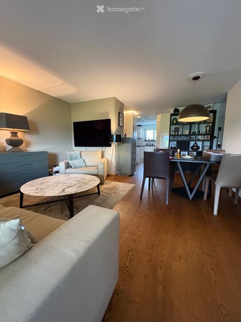 Open-plan living and dining area with hardwood flooring, a white sofa, a round coffee table, a dining table with chairs, and a TV mounted on the wall.