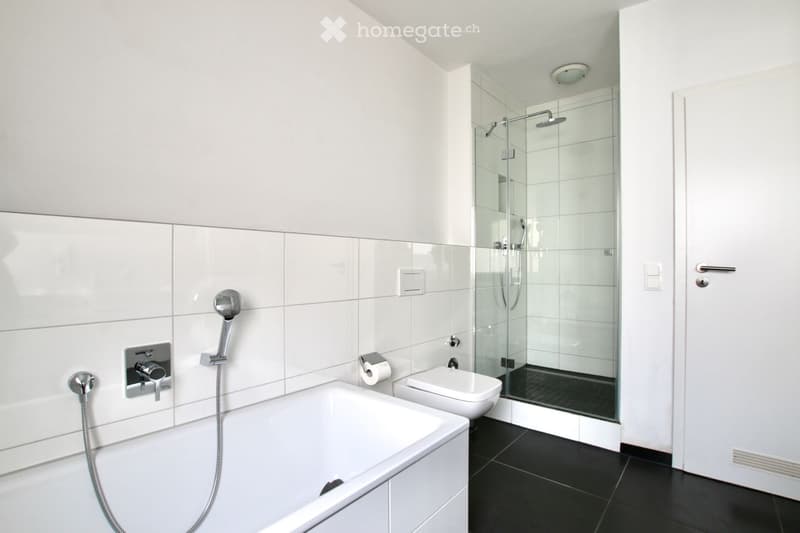Modern bathroom with a bathtub, toilet, and glass-enclosed shower. Features white tiles, black flooring, and sleek fixtures.