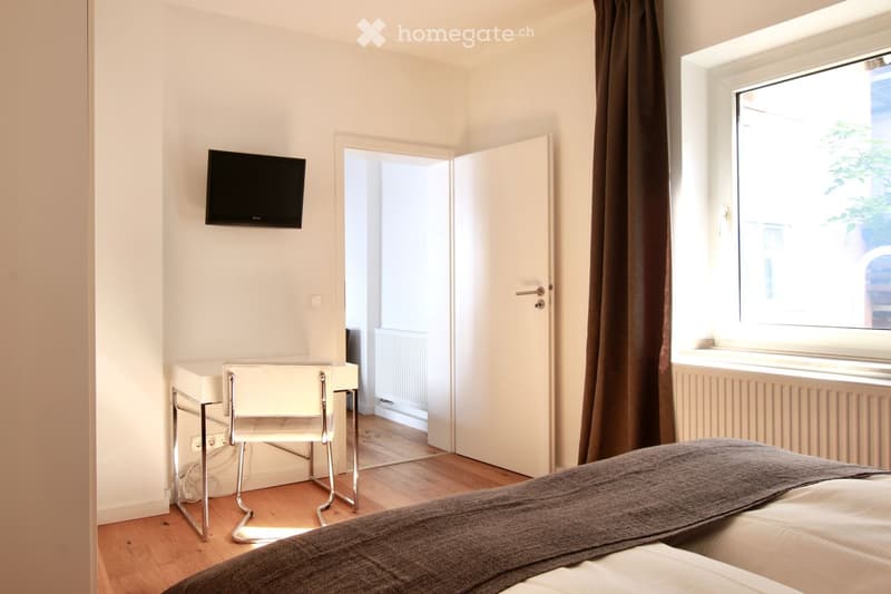 Cozy bedroom with a bed, a chair, a small table, and a wall-mounted TV. The room features wooden flooring and large windows with curtains.