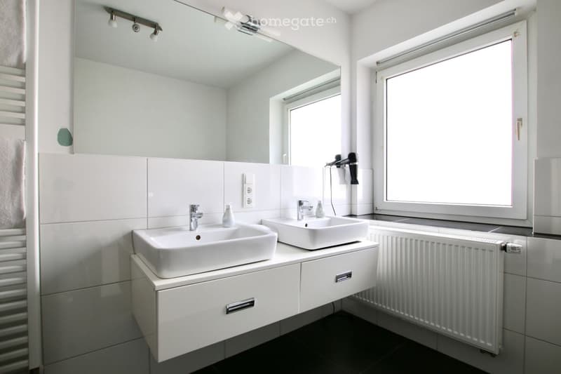 Modern bathroom with two white sinks, chrome faucets, a large mirror, a radiator, and a towel on the side.