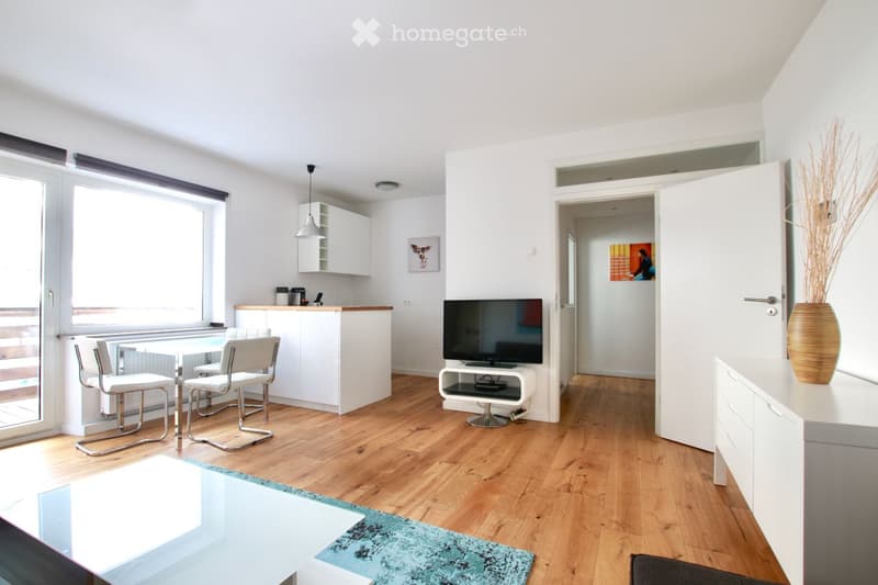 Modern living room with wooden flooring, white walls, and large windows. Features a dining table with chairs, a television on a stand, a cabinet, and a decorative vase with dried plants.