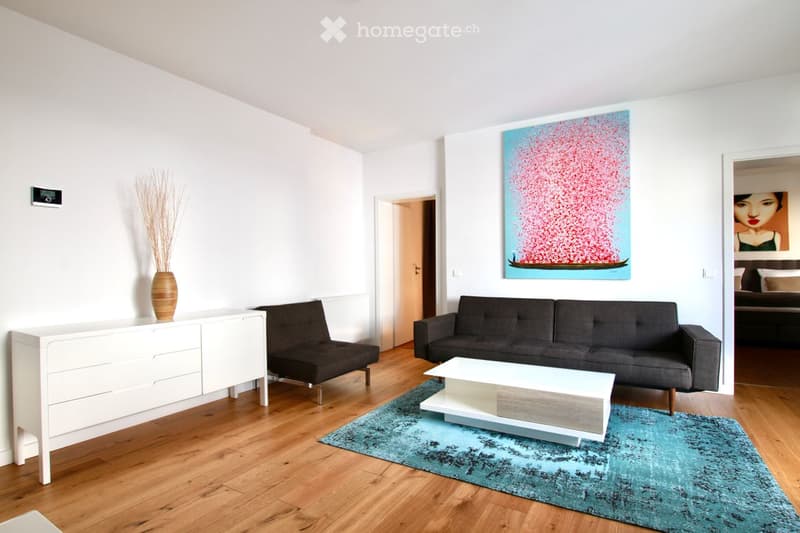 Modern living room with light wooden flooring, white walls, grey sofa, white coffee table, small black chair, white cabinet, decorative vase, and artwork on the wall.